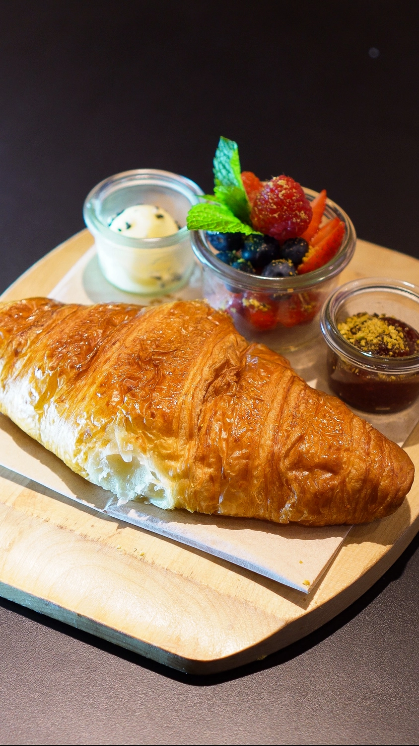 NOA - Café Brunch Konzept - Ein blättriges, goldbraunes Croissant, serviert auf einem Holzbrett mit Beilagen.